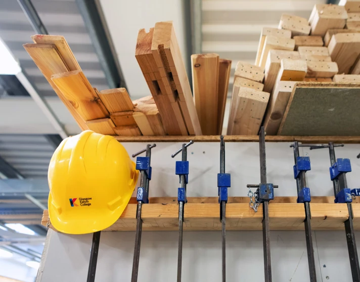 Woodworking equipment including clamps, timber planks, and a bright yellow safety helmet neatly arranged on a storage rack in a joinery workshop. Woodworking equipment including clamps, timber planks, and a bright yellow safety helmet neatly arranged on a storage rack in a joinery workshop.